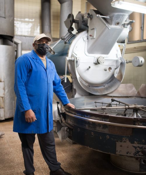 Mid adult coffee roaster standing next to coffee roasting machine. Interior of coffee production workshop with set up roasting equipment. Person in uniform and mask. Production, food concept